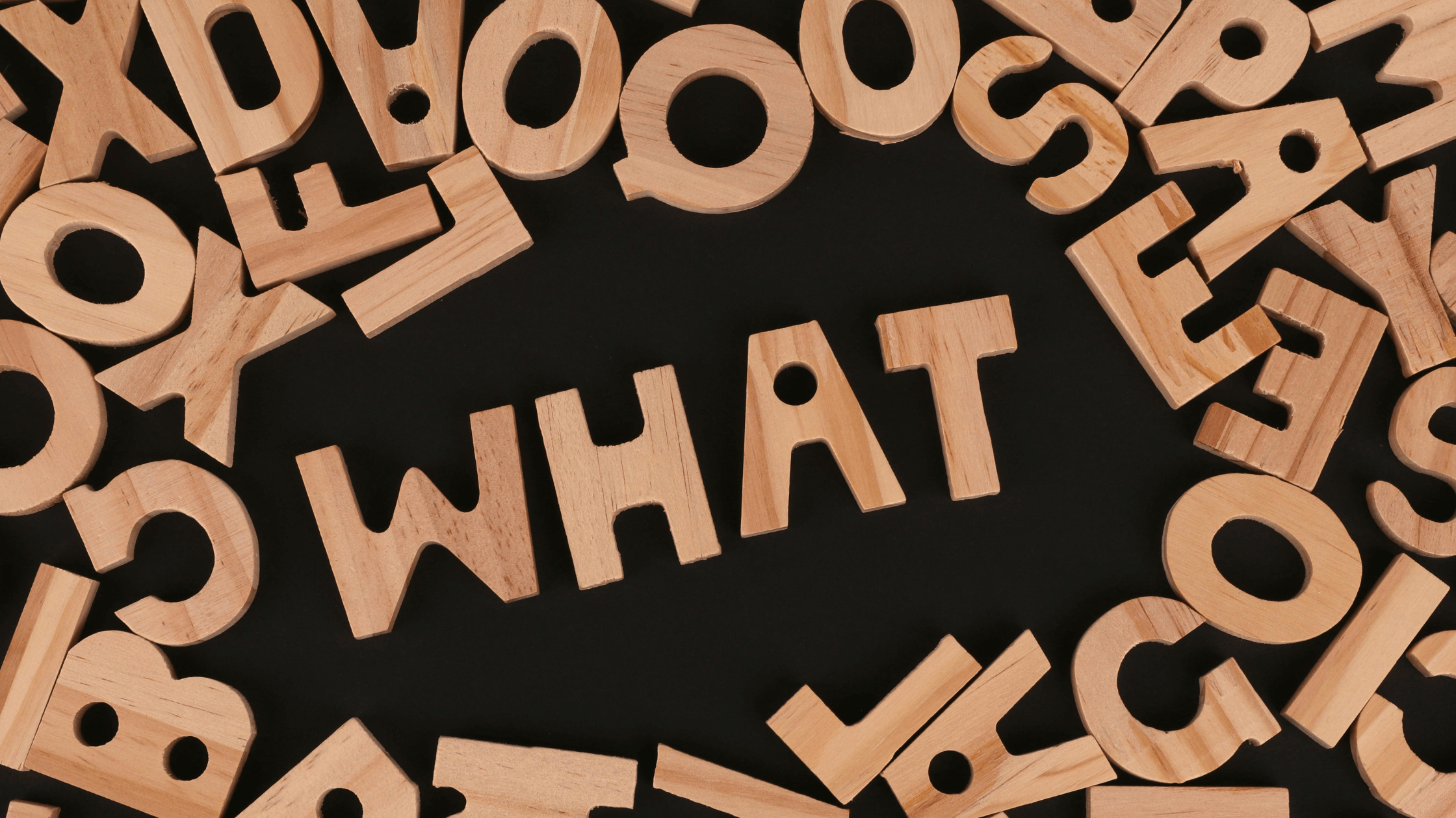 Wooden letters arranged on a black background forming the word “WHAT.”