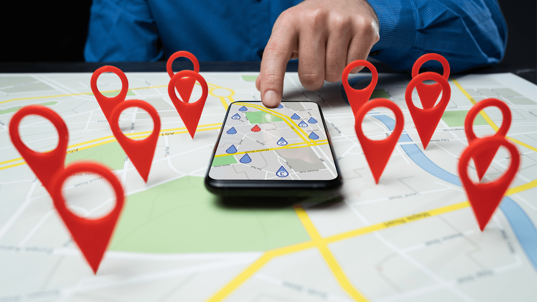 Person pointing at a smartphone placed on a paper map with red location markers surrounding it, symbolizing GPS navigation and multiple locations.