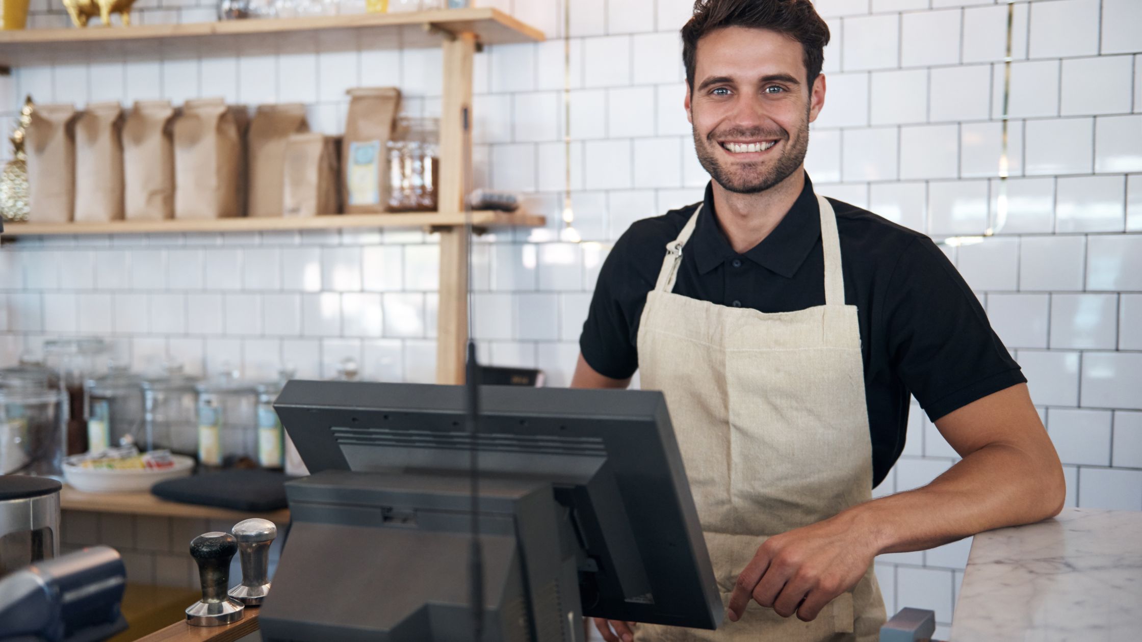 Man wearing apron standing behind a POS terminal in a café, smiling at the camera.