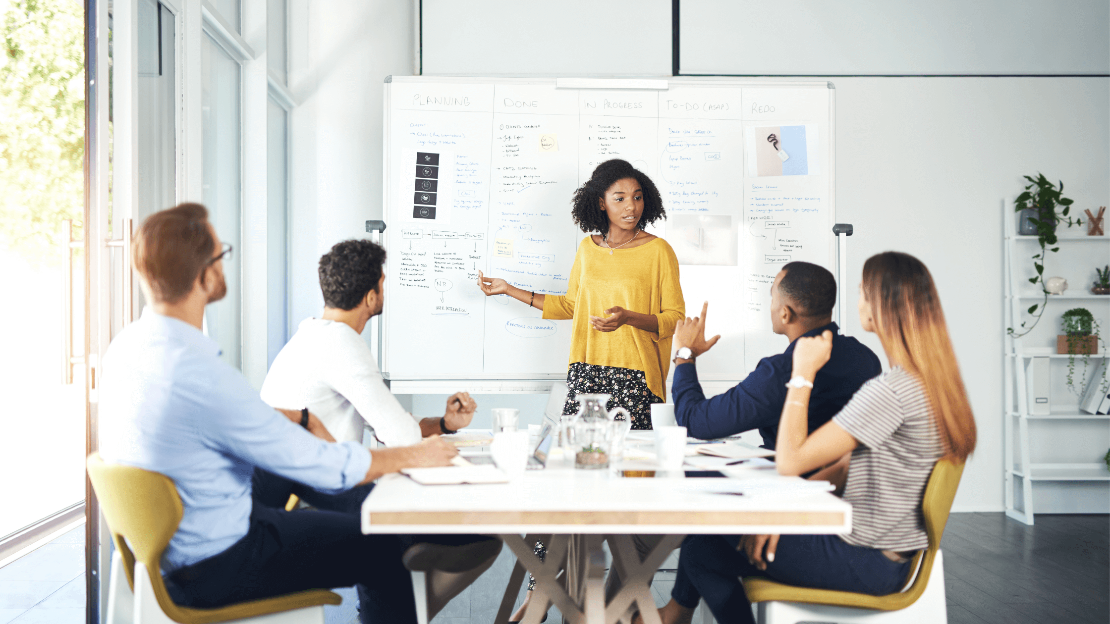 Woman leading a business meeting, pointing to notes on a whiteboard while team members listen and discuss around a conference table.